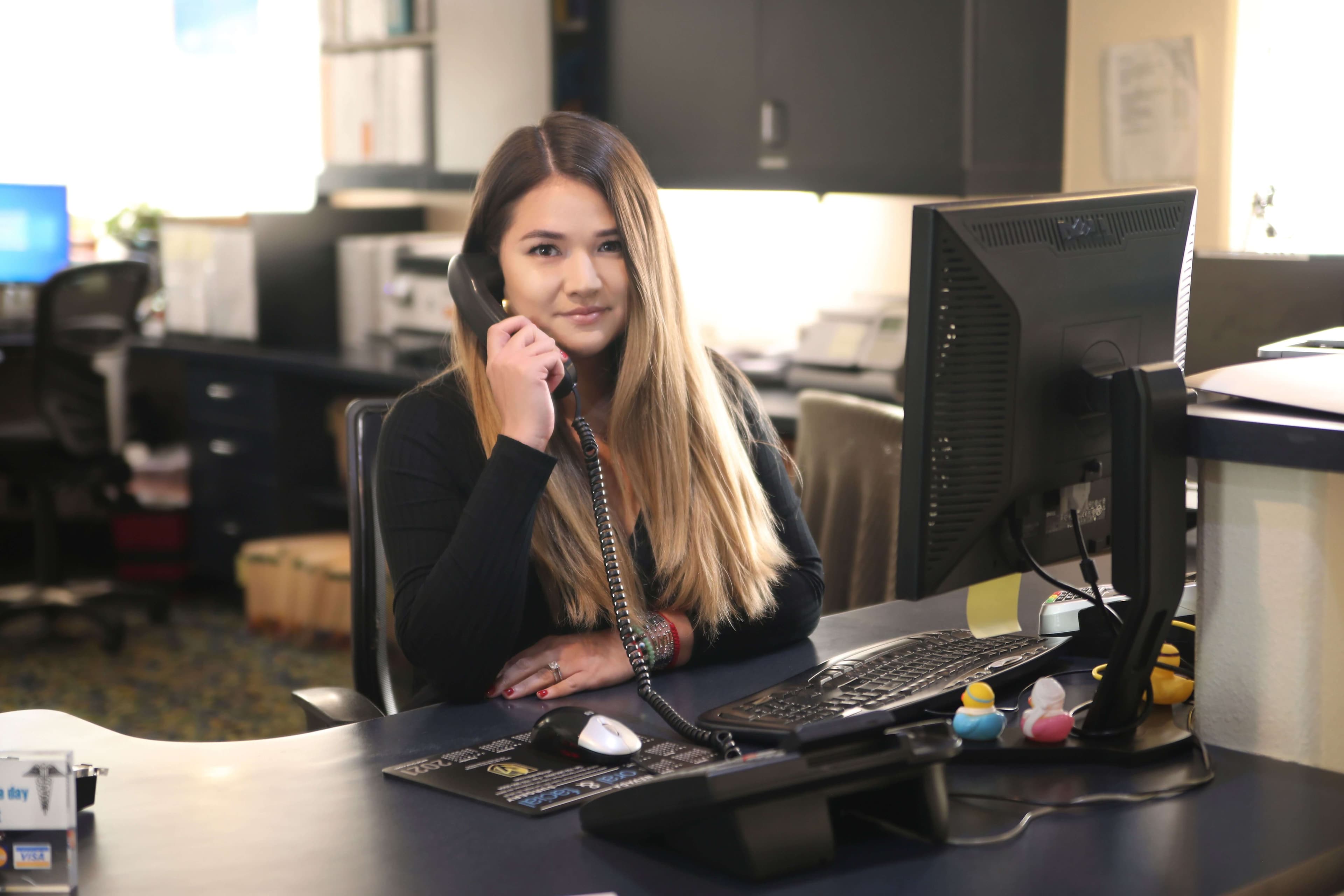 Blonde woman sitting in front desc, smiling, holding a phone, welcoming people to contact an office